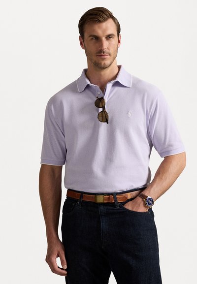 Helllila Polohemd mit Kragen und kurzen Ärmeln, kombiniert mit dunkelblauen Jeans und einem braunen Gürtel. Sonnenbrillen liegen auf dem Kragen.