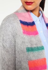 Cardigan en tricot gris avec des rayures multicolores dans les tons rose, bleu, orange et vert. Texture douce et design ample avec un devant ouvert.