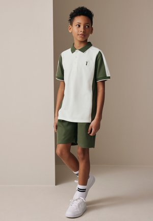 Junge steht mit entspannten Händen, trägt weißes und grünes Poloshirt, grüne Shorts, weiße Sneakers und weiße Socken mit schwarzen Streifen.