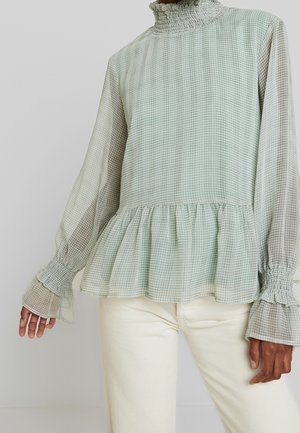 Woman wearing a light green and white checkered blouse with ruffled high neck, flared cuffs, peplum hem, and white pants.