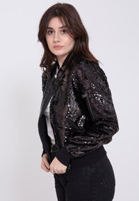 Czarna kurtka bomberka wykonana z materiału pokrytego cekinami o błyszczącej fakturze, z prążkowanymi mankietami i zamknięciem na zamek z przodu. Dodatkowe szczegóły to boczne kieszenie.