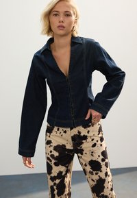 Chaqueta vaquera con escote en V profundo y cierre de botones, combinada con pantalones beige con un patrón de manchas negras. Texturas suaves y diseño ajustado.