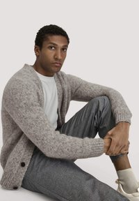 Lichtbruine gebreide cardigan met geribbelde manchetten, gecombineerd met grijze broek. Model zit en toont de zachte textuur en de knoopdetails op de trui.