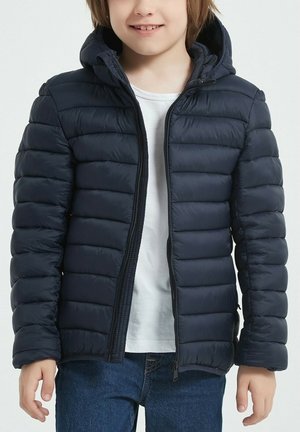 Veste matelassée bleu marine avec matelassage horizontal, fermeture éclair, capuche et deux poches latérales. Portée sur un t-shirt blanc et un jean bleu.