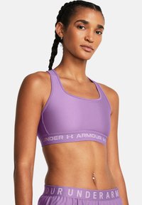 Trenings-BH i lys lilla stoff, med en bred elastisk kant som har "UNDER ARMOUR"-logoen. Glatt, tettsittende design med åpen rygg.