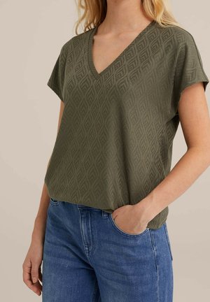Oliwkowa bluzka z krótkim rękawem w serek z teksturowanym wzorem liści, zestawiona z niebieskimi dżinsami. Materiał wydaje się miękki i lekki.