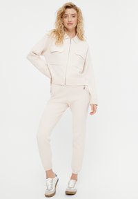 Giacca rosa chiaro con zip e due tasche sul petto, abbinata a pantaloni jogger coordinati. Texture a maglia morbida, con cerniere e design aderente.