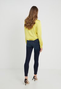 Dlouhý rukáv žluté hedvábné blůzy v kombinaci s tmavými skinny džínami, s jednoduchým designem a těsným střihem. Doplňují ji béžové lodičky.