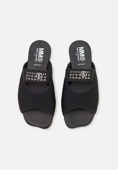 Sorte slipsandaler laget av myk tekstil med runde tær og en bred stropp med nummerert trykk. Sålen er glatt og flat.