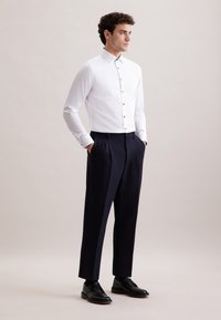 Witte button-up shirt met een puntige kraag en donkere knopen, gecombineerd met marineblauwe geplooide broek en zwarte veterschoenen. Gladde texturen.