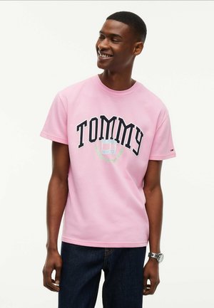 Jonge man met een roze Tommy Jeans T-shirt en donkere spijkerbroek, glimlachend en naar de zijkant kijkend met een horloge om zijn linkerpols.
