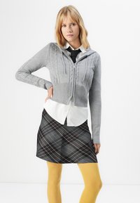 Siva zip-up pulover z okrasno pletenico, prekrit nad belim srajčnim ovratnikom in krilom s črno-sivim karo vzorcem. Rumene nogavice dopolnjujejo videz.