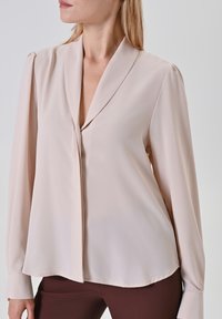 Helle beigefarbene Bluse mit einem Schalkragen, langen Ärmeln und einer glatten Textur; verfügt über eine Vorderblende und eine entspannte Silhouette.