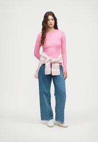 Dugih rukava ružičasti rebrasti top, zavezan svijetloružičasti i bijeli prugasti džemper oko struka, široke plave traperice i bijele tenisice.