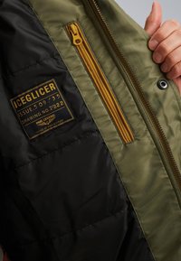 Vadderad jacka med en olivgrön och svart färgskala, med en framträdande gul dragkedja och en etikett som säger "ICEGLICER".