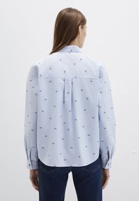 Light blue, long-sleeved shirt with floral embroidery, back pleat detail, rounded hem, and button cuffs, made of soft fabric.