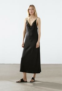 Abito in satin nero con scollatura a V rifinita in pizzo, dotato di spalline sottili e regolabili e design a lunghezza completa, abbinato a sandali neri con tacco basso.