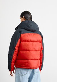 Rød og sort pufferjakke med hætte, der har et kviltet design og en blød tekstur. Synlige sømme langs ærmerne og kanten.