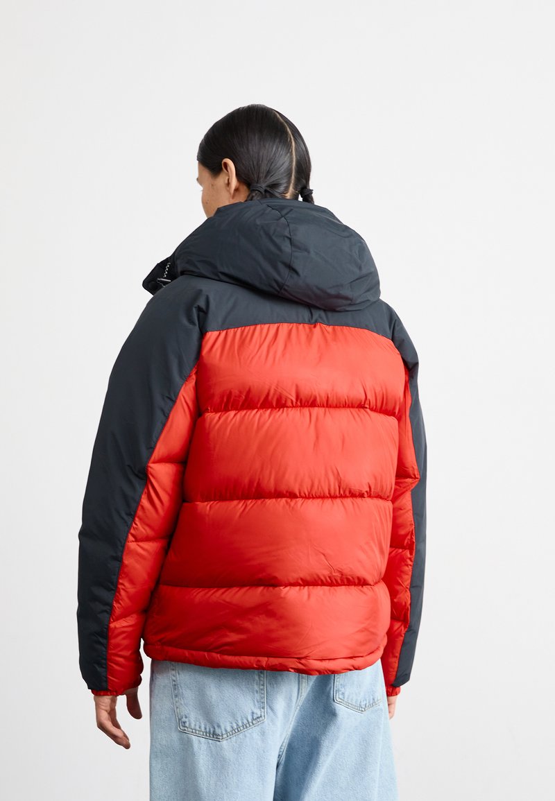 Rote und schwarze Steppjacke mit Kapuze, die ein gestepptes Design und eine weiche Textur aufweist. Sichtbare Nähte entlang der Ärmel und des Saums.