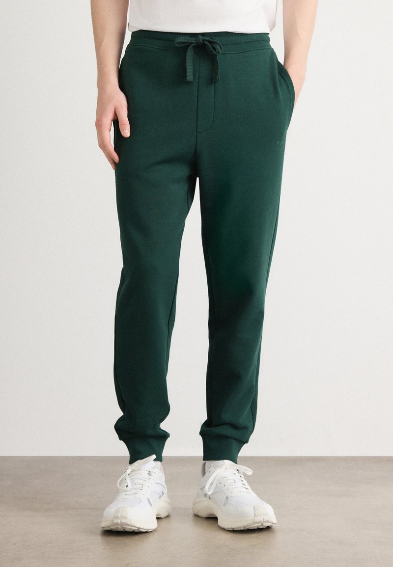 Joggers verdi in cotone misto con vita elastica, cordoncino regolabile, tasche laterali e polsini a costine, abbinati a sneakers bianche.