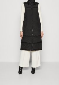 Svart pufferväst med hög krage, med en framdragkedja, två sidofickor och en quiltad textur. Buren över vita långärmade tröjor och vida byxor.