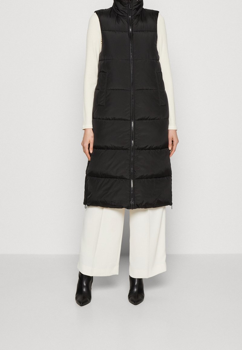 Svart pufferväst med hög krage, med en framdragkedja, två sidofickor och en quiltad textur. Buren över vita långärmade tröjor och vida byxor.