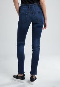 Jeans ajustados de denim azul oscuro con cinco bolsillos, una altura media y una textura sutil. Usados con zapatillas negras sin cordones.