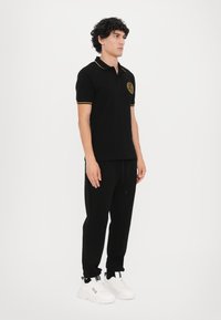 Cămașă polo neagră cu accente galbene și logo, combinată cu pantaloni jogger negri și adidași albi. Modelul stă într-o poziție neutră pe un fundal alb.