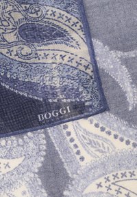 Mėlyna raštuota skara, pagaminta iš lengvos medžiagos. Puikuojasi paisley dizainu ir nertos etiketės su prekės ženklo pavadinimu "BOGGI" balta spalva.