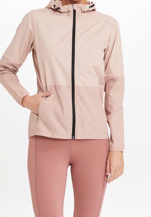 Frau trägt eine hellrosa Kapuzenjacke mit Reißverschluss, eine Hand in der Tasche und mauvefarbene Leggings vor weißem Hintergrund.