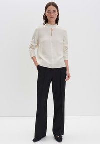 Donna in piedi con le mani in tasca, che indossa un top a maniche lunghe in crema con dettagli a chiave e pantaloni neri a gamba larga.