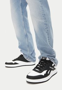 Osoba w jasnoniebieskich dżinsach i czarno-białych trampkach Reebok stoi na białym tle.