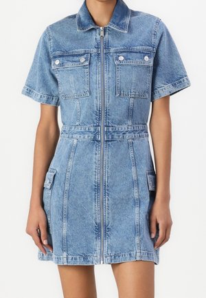 Kvinde iført kortærmet lyseblå denimkjole med lynlås foran, to brystlommer med klap og to sidelommer med klap.