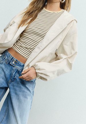 Femme portant une veste beige zippée sur un crop top rayé crème et noir avec un jean bleu taille haute sur un fond clair.