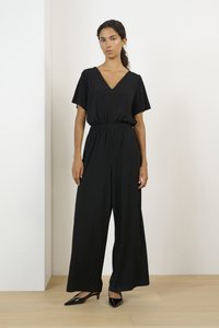 Svart jumpsuit med korta ärmar, V-ringning, elastisk midja och vida ben. Modellen bär svarta pumps med spetsiga tår och står på ett trägolv.