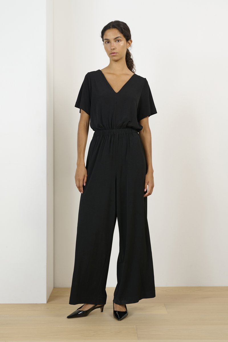 Svart jumpsuit med korta ärmar, V-ringning, elastisk midja och vida ben. Modellen bär svarta pumps med spetsiga tår och står på ett trägolv.