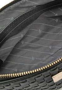 Interior de bolso tejido negro con bolsillo con cremallera, forrado en tela gris con patrón que presenta sutiles rayas y símbolos. Detalles de cremallera dorados.