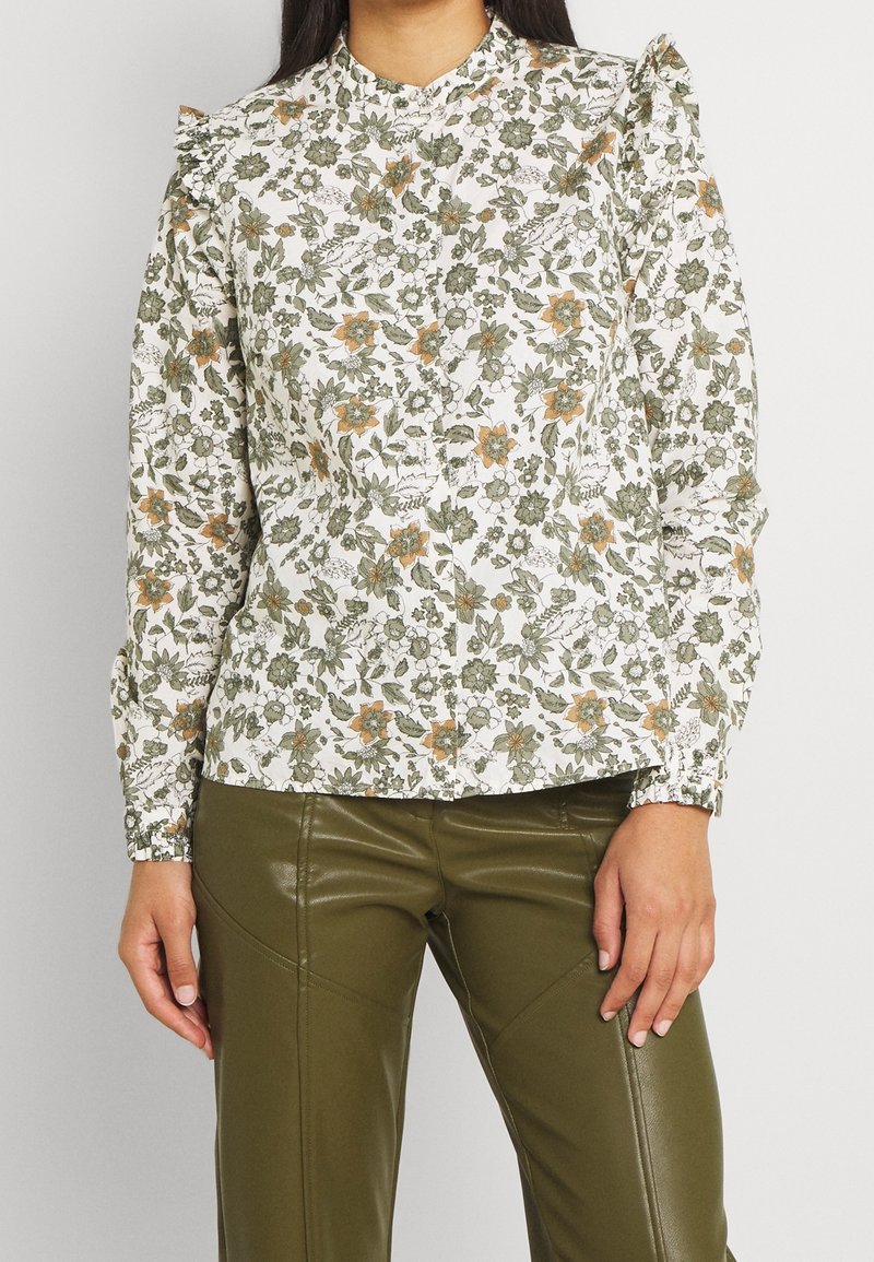 Blouse blanche à motifs floraux verts et orange, tissu léger, col montant et poignets élastiques. Associée à un pantalon ajusté vert olive.