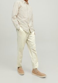 Man wearing beige striped long-sleeve shirt, cream-colored loose pants, beige socks, and tan slip-on shoes standing with hands in pockets.