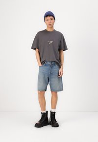 Siva oversize majica s tiskanim besedilom, modri jeans kratke hlače in črne čipkaste čevlje. Model nosi modro pleteno kapo in bele nogavice.