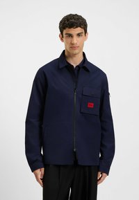 Navy blue jacket made of a smooth fabric, featuring a front zip, large chest pocket with red label, and a classic collar design.