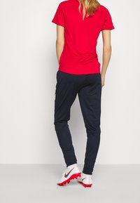 Röd sportig kortärmad tröja, marinblå tajta byxor och vita fotbollsskor med röda accenter. Slät tyg med strömlinjeformat mönster.