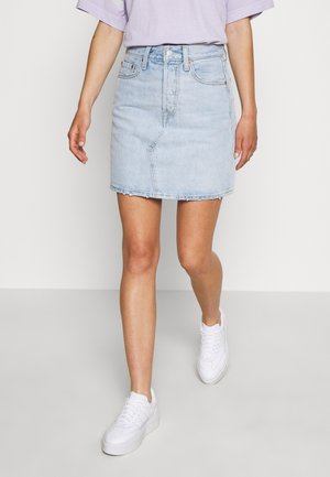Frau trägt einen hellblauen Jeansrock mit rohem Saum, weiße Sneakers und ein hellviolettes Kurzarmshirt, läuft auf einem schlichten Boden.