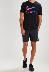Camiseta negra de Nike de manga corta con texto colorido "RUNNING" y un logo; combinada con pantalones cortos oscuros y zapatillas deportivas negras.