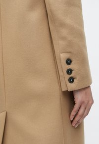 Μανίκι από μπεζ μάλλινο παλτό με τρία μαύρα κουμπιά στη μανσέτα και ένα χαλαρό χέρι τοποθετημένο δίπλα του.