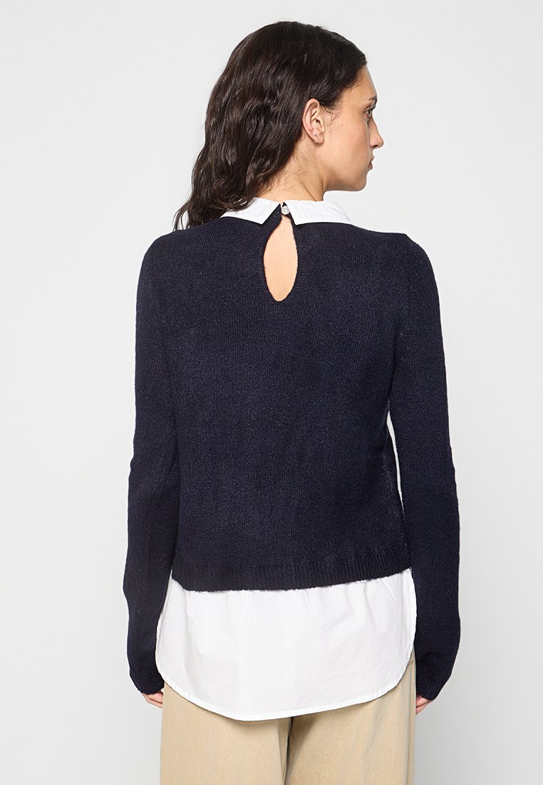 Pull marin en tissu tricoté doux, avec des manches longues et un détail en forme de trou de serrure au dos, porté par-dessus une chemise blanche.