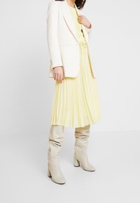 Blazer beige su un vestito plissettato giallo chiaro, abbinato a stivali crema al ginocchio. Texture morbide e elementi di design eleganti.