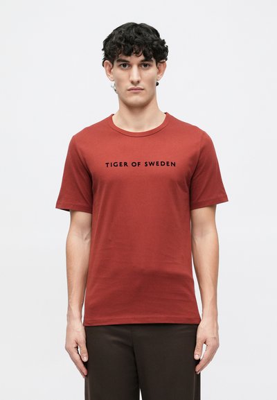 T-shirt rouge à manches courtes en coton avec un col rond, comportant le texte "TIGER OF SWEDEN" imprimé en noir sur la poitrine.