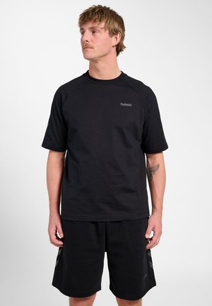 Mand med kort hår iført sort hummel t-shirt og sorte shorts, stående og kigger let til venstre, viser underarms-tatovering.