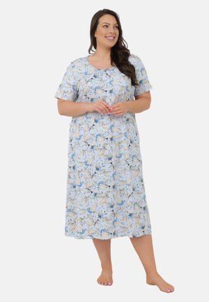 Kobieta w midi sukience z krótkim rękawem w niebiesko-beżowy kwiatowy wzór, stojąca boso, z rękami splecionymi, uśmiechająca się i patrząca na bok.